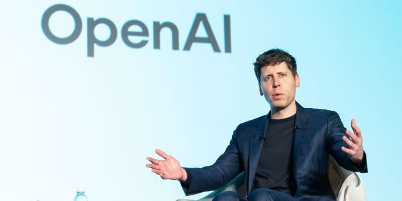 Sam Altman speaking on stage during an OpenAI event with the OpenAI logo displayed in the background.